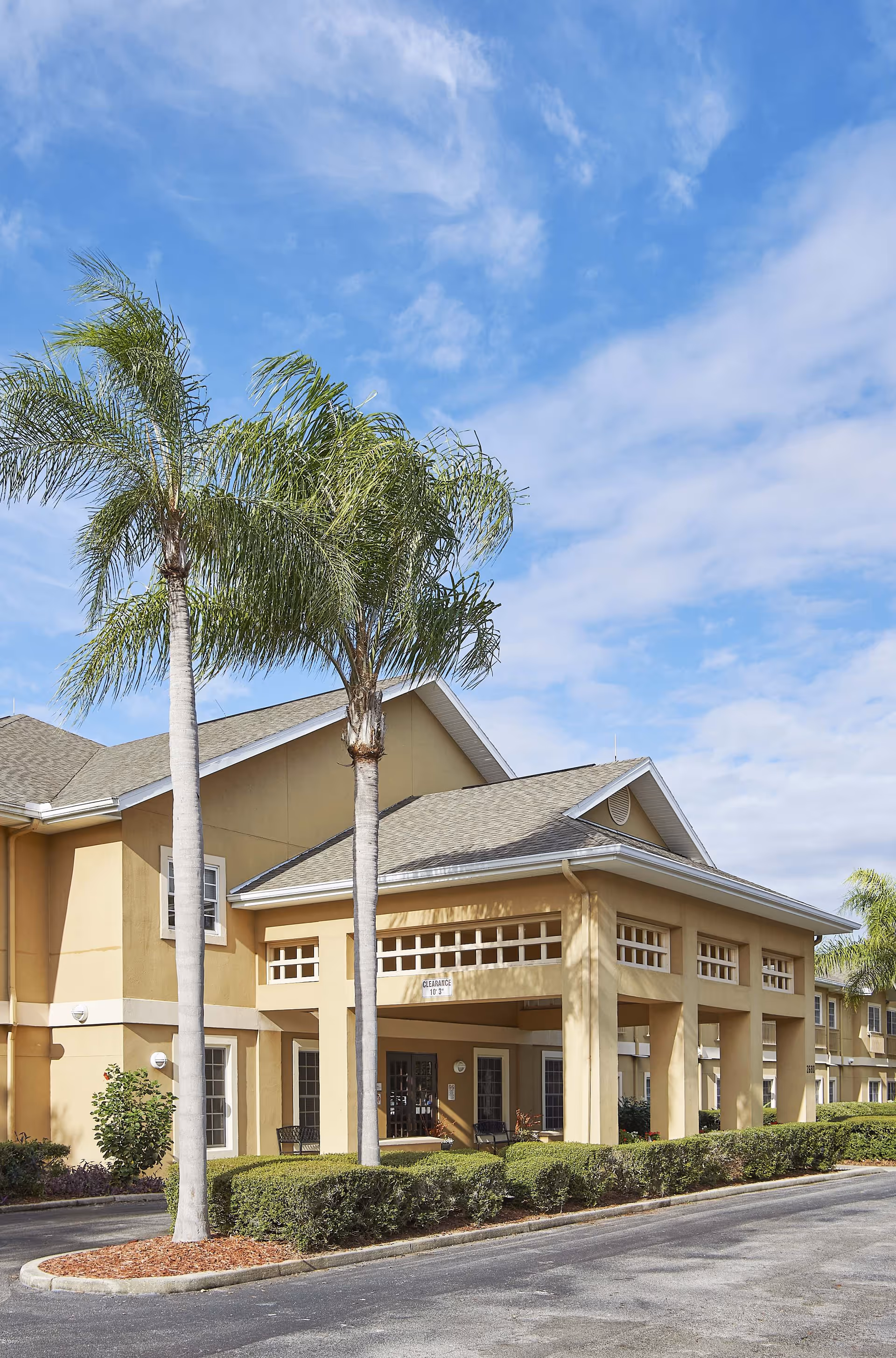 Front entrance of a beige senior living building with palm trees and a covered porte-cochère under a blue sky.