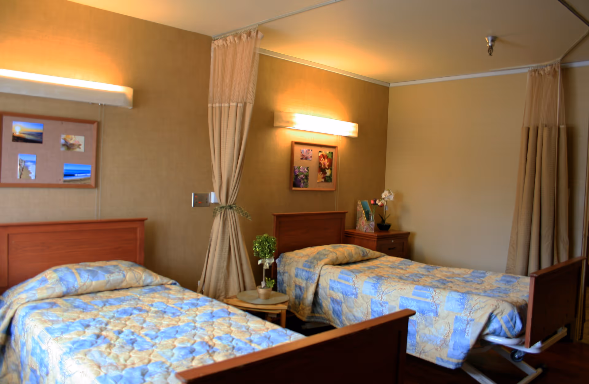 A shared patient bedroom with two twin beds, a bedside table, wall lights, and privacy curtains.