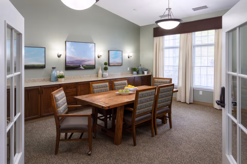A dining room with a wooden table surrounded by six chairs with patterned upholstery. The room has light green walls, three landscape paintings, and a long countertop with cabinets underneath. Large windows with white blinds and cream-colored curtains allow natural light to fill the space. A ceiling light fixture hangs above the table.