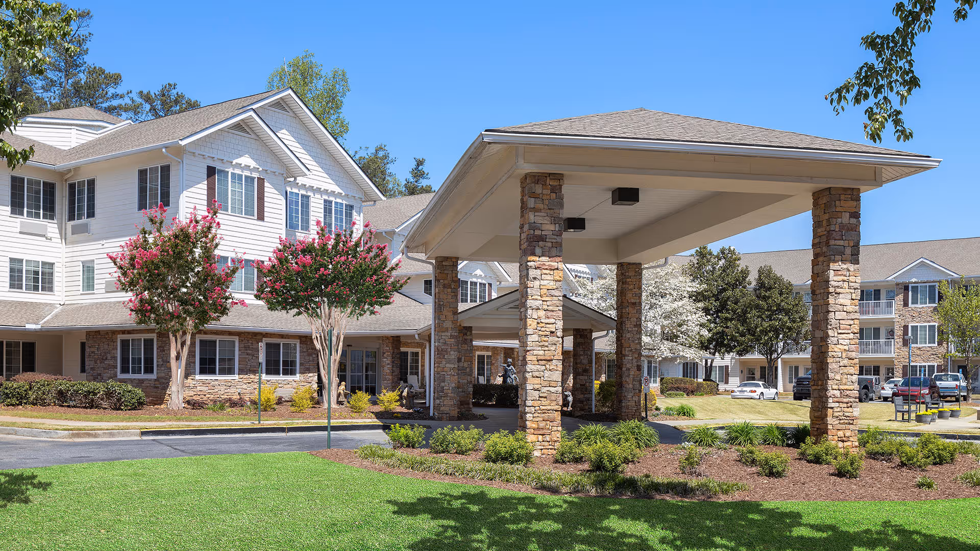 Exterior view of Laurel Grove by Barclay House, showing a multi-story building with white siding and stone accents. The entrance features a covered drop-off area supported by stone pillars. The surrounding landscape includes green grass, flowering trees, and shrubs under a clear blue sky.