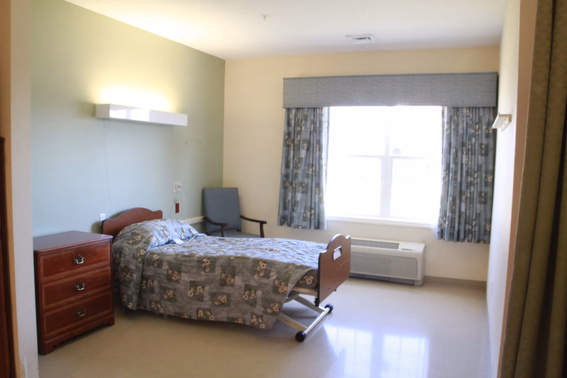 A simple, clean bedroom in a senior living facility with a single bed covered in a floral patterned quilt, a wooden nightstand with three drawers, a gray armchair, a large window with blue floral curtains, and a wall-mounted light fixture above the bed.