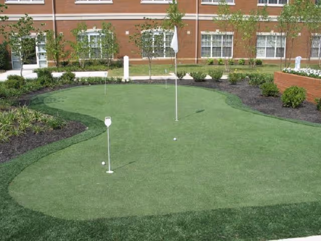 Outdoor putting green with flags and golf balls in a landscaped courtyard in front of a brick building.