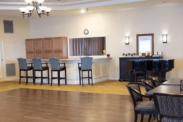 Interior view of a senior living facility lounge area with a bar counter and four high chairs. There is a black grand piano against the wall with decorative items on top, and a large mirror above the bar. Several dining tables and chairs are partially visible on the right side. The room has warm lighting and a combination of carpet and wooden flooring.