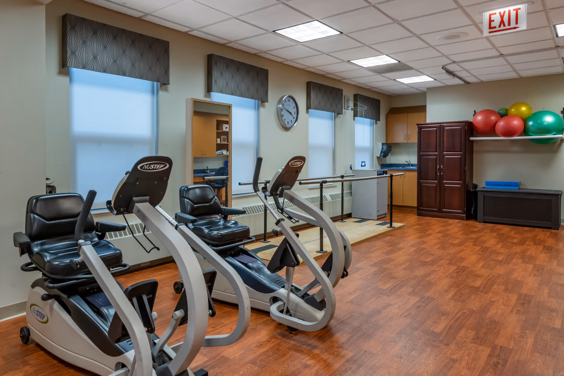 A fitness room with two recumbent exercise bikes, wooden flooring, and a wall-mounted handrail for support. The room has three windows with patterned valances, a clock on the wall, a wooden cabinet, a shelf holding colorful exercise balls, and a small kitchenette area in the back.