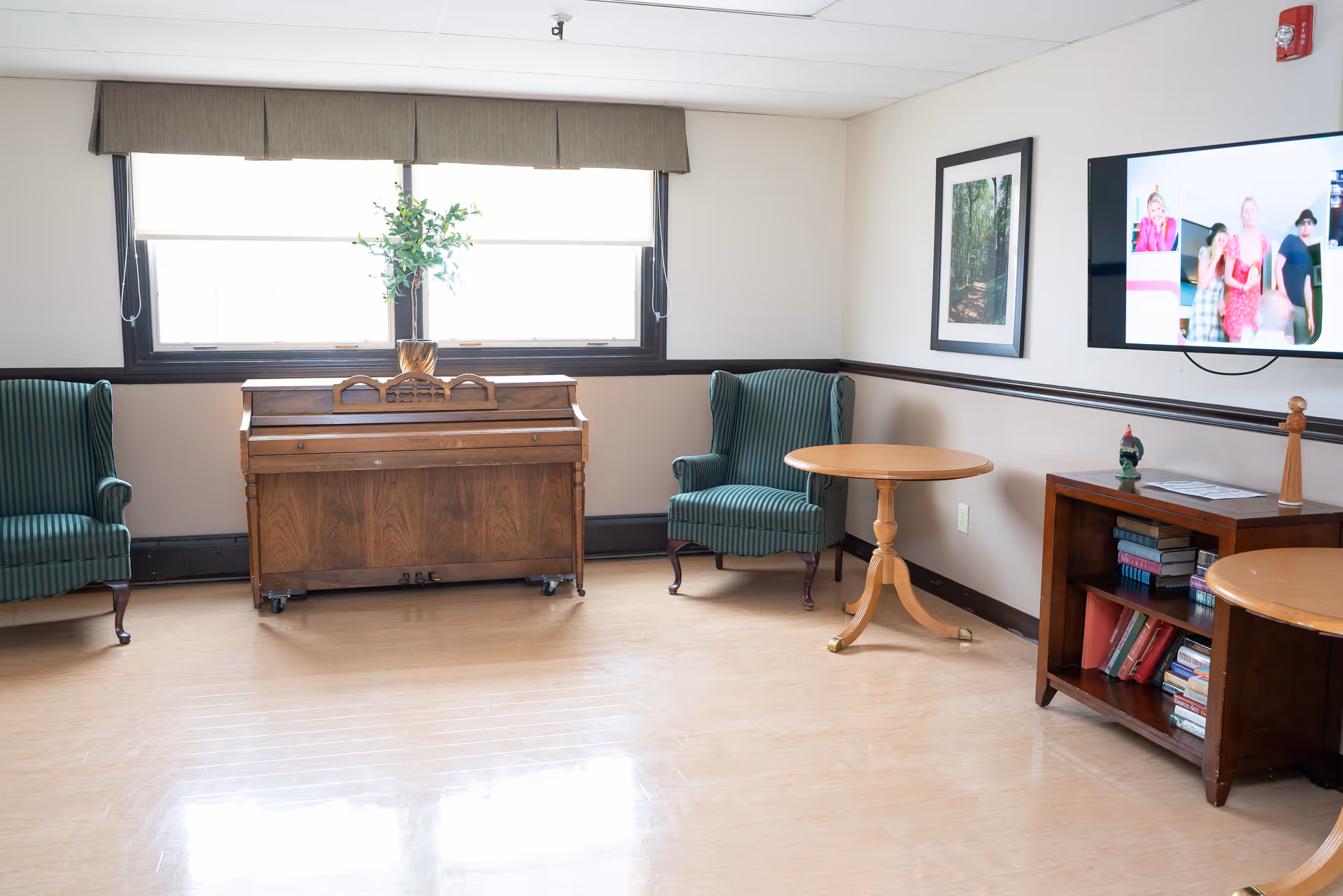 A bright room with two green striped armchairs, a wooden piano with a plant on top, two round wooden tables, a wooden bookshelf filled with books, a framed picture on the wall, and a flat-screen TV displaying a group of people.