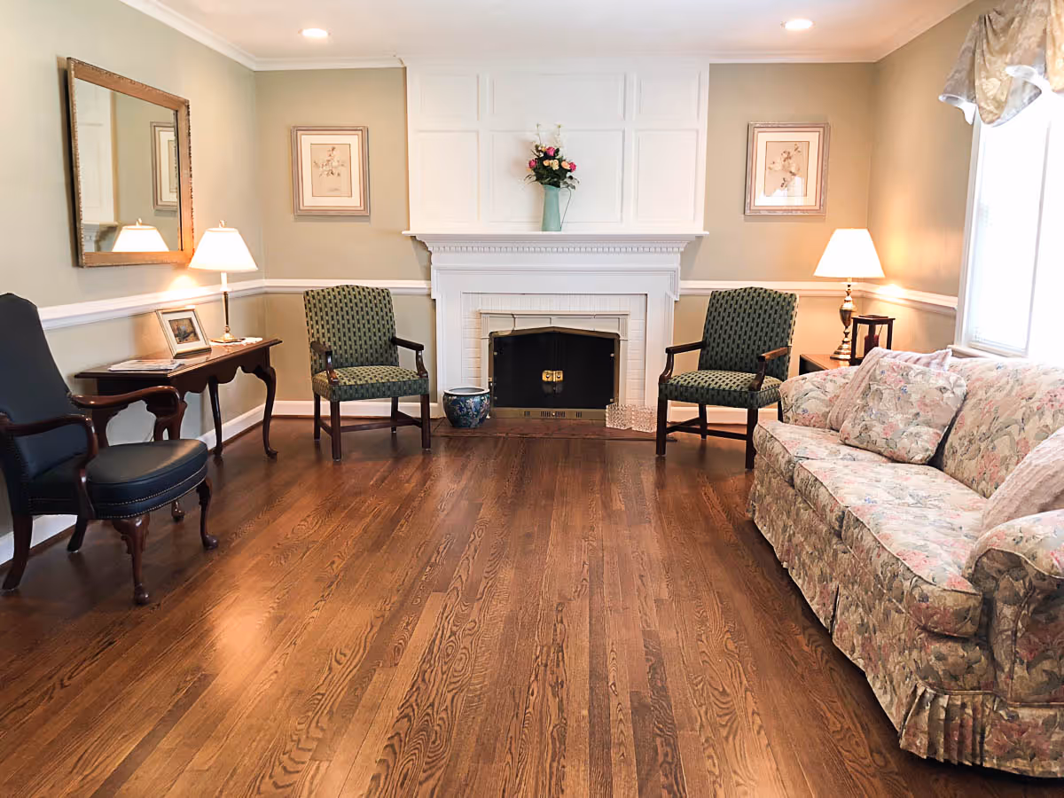 A cozy living room with wooden floors, a floral patterned sofa on the right, two green upholstered chairs facing a white fireplace with a vase of flowers on the mantel. There are two table lamps on either side of the room, a large mirror on the left wall, and framed artwork above the fireplace and chairs.