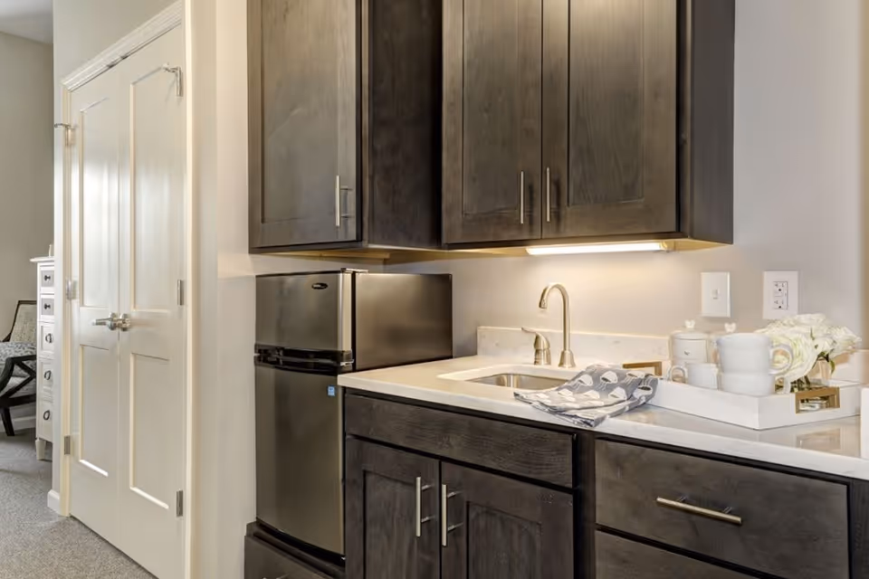 A small kitchenette area with dark wood cabinets, a stainless steel mini refrigerator, a sink with a modern faucet, and a white countertop. On the countertop, there is a white tray holding white cups, a teapot, and a small flower arrangement. A double door closet is visible to the left, and part of a chair and dresser can be seen in an adjacent room.
