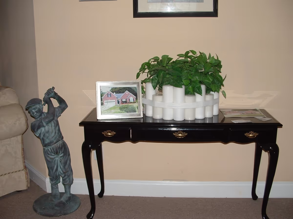 A black wooden console table with three drawers, placed against a beige wall. On the table, there is a framed photo of a house, a white decorative planter with green leafy plants, and some papers. To the left of the table, there is a statue of a boy in vintage golf attire holding a golf club in a swinging pose. Part of a beige upholstered chair is visible on the far left.