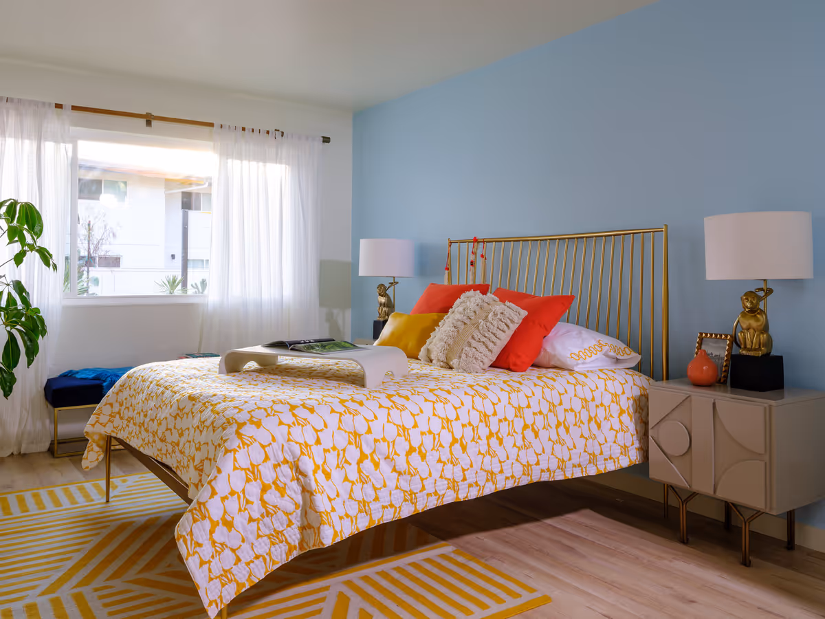 Bright modern bedroom with a gold metal bedframe, yellow-and-white patterned bedding, colorful pillows, bedside tables with lamps, and a large window with sheer curtains.
