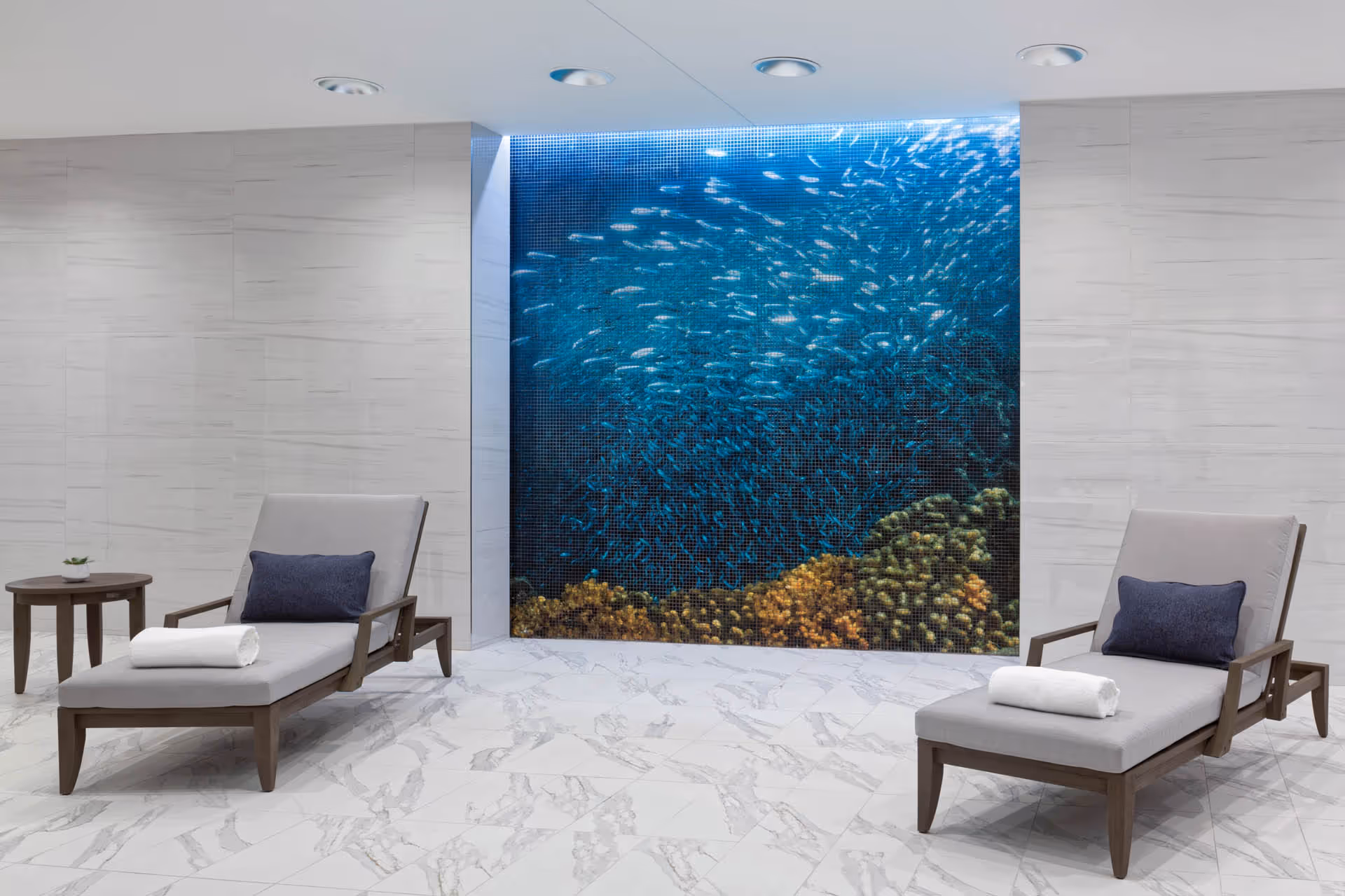 Two chaise lounges with pillows and rolled towels in a marble-floored relaxation room facing a large illuminated mosaic of an underwater coral and fish scene.