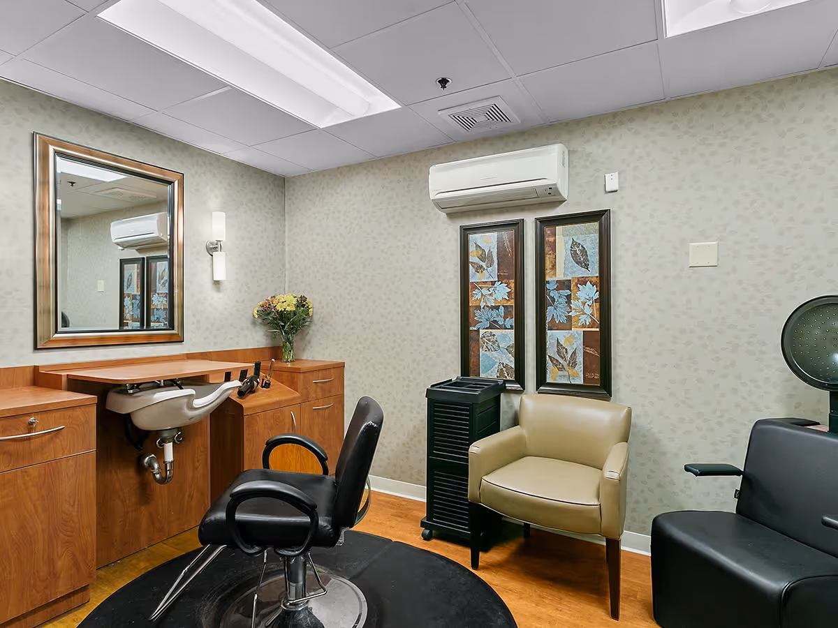 Interior of a hair salon area in a senior living facility with a black salon chair in front of a wooden vanity with a sink, a large mirror, and a wall-mounted light. There is a beige armchair, a black salon dryer chair, two framed leaf-themed artworks on the wall, and an air conditioning unit above them.