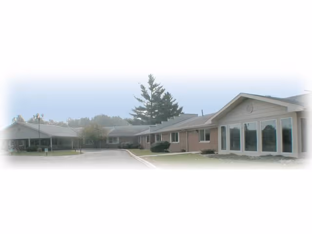Exterior view of a single-story senior living facility building with large windows and a driveway in front, surrounded by some trees and bushes under a clear sky.