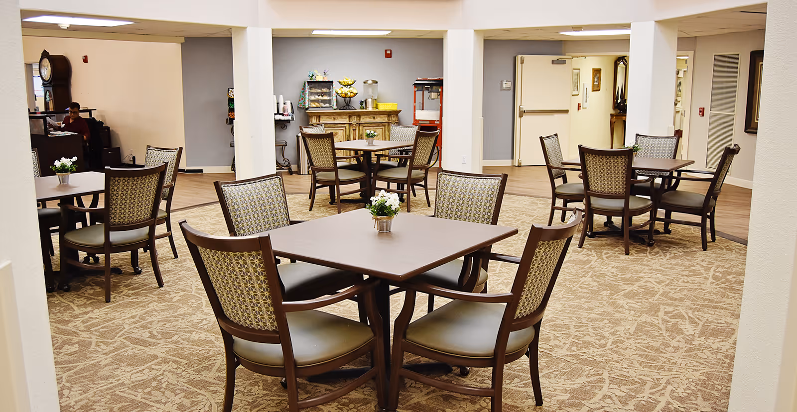 A dining area in a senior living facility with several square tables, each surrounded by four cushioned chairs with patterned backs. Small flower arrangements are placed on each table. The room has beige patterned carpet and light-colored walls. In the background, there is a wooden cabinet with a water dispenser, a popcorn machine, and some cups. A person is seated at a desk in the far left corner.