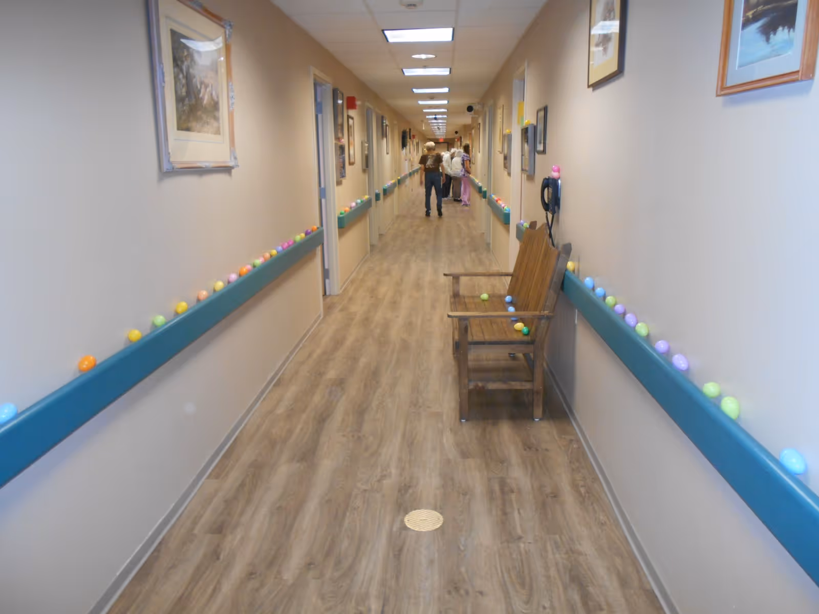 Long hallway in a senior living facility with wood flooring and beige walls. The hallway has teal handrails decorated with colorful plastic Easter eggs. There is a wooden bench on the right side and framed pictures hanging on the walls. Two people are visible walking down the hallway in the distance.