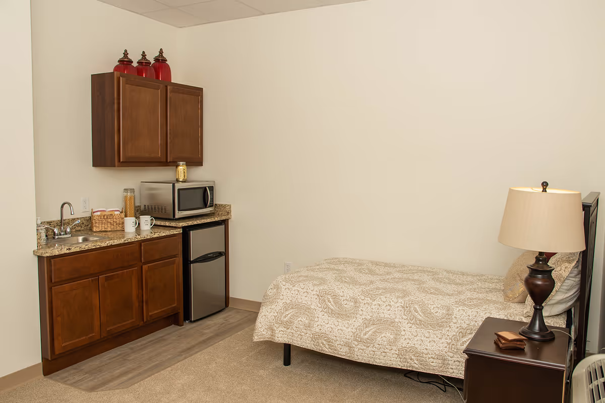A small room with a single bed covered in a beige patterned bedspread, a wooden nightstand with a lamp, and a kitchenette area with wooden cabinets, a small sink, a microwave, a mini refrigerator, and some jars and mugs on the countertop.