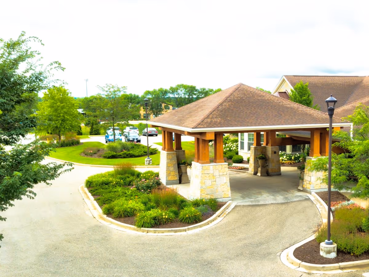 Entrance area of The Auberge at Brookfield featuring a covered driveway with stone pillars and wooden beams, surrounded by landscaped greenery and a few parked cars in the background.