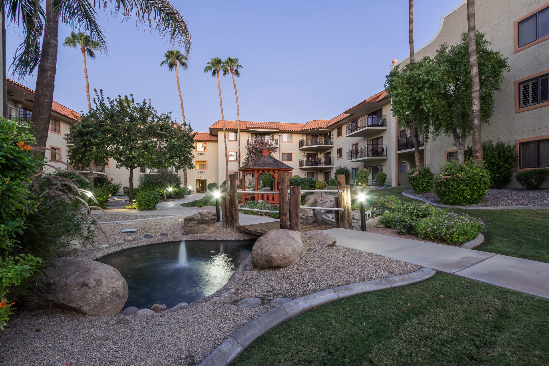 Outdoor courtyard area of a senior living facility with a small pond featuring a fountain, a wooden footbridge, a red gazebo, palm trees, and surrounding three-story buildings with balconies during dusk.