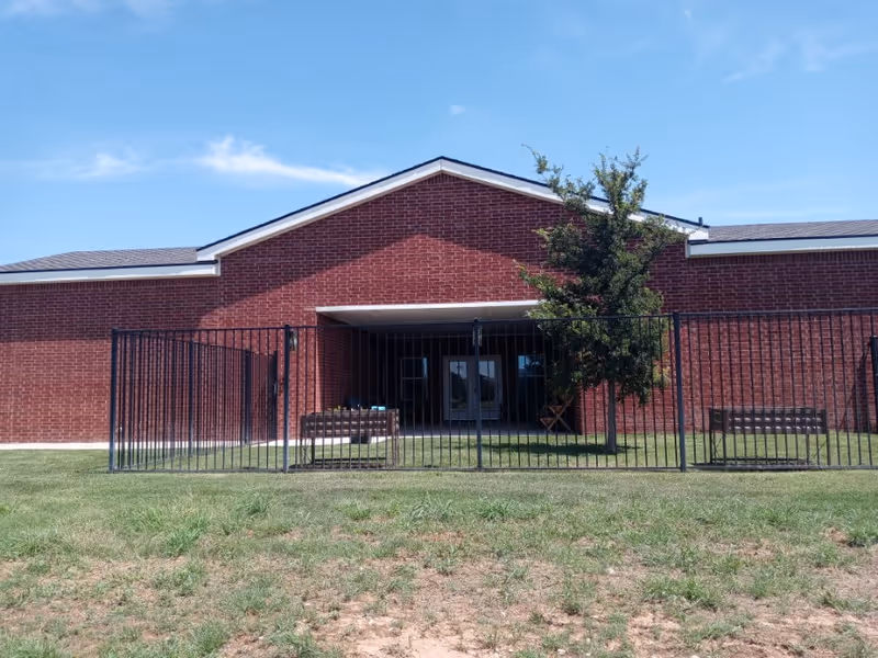 Exterior view of a single-story red brick building with a gabled roof under a clear blue sky. In front of the building is a black metal fence enclosing a small grassy area with two benches and a tree. The building has a covered entrance with glass doors.