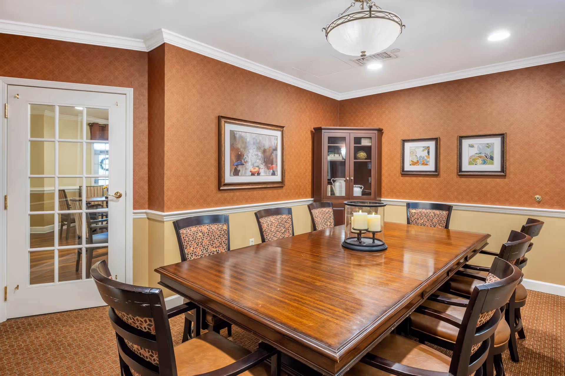 A formal dining room with a long polished wooden table surrounded by chairs, wall art, and a glass-front cabinet.