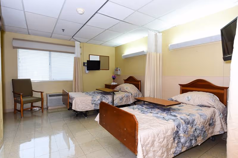 A rehabilitation facility room with two hospital-style beds, each with floral patterned bedspreads and overbed tables. The room has beige walls, a window with blinds, a green armchair, a wall-mounted TV, and privacy curtains between the beds.
