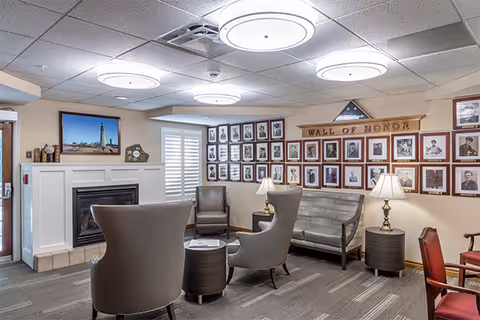 A cozy sitting area in an assisted living facility featuring a fireplace, several chairs arranged around small tables, a bench, and a wall displaying framed photographs labeled 'Wall of Honor'. The room has a carpeted floor, ceiling lights, and window shutters.