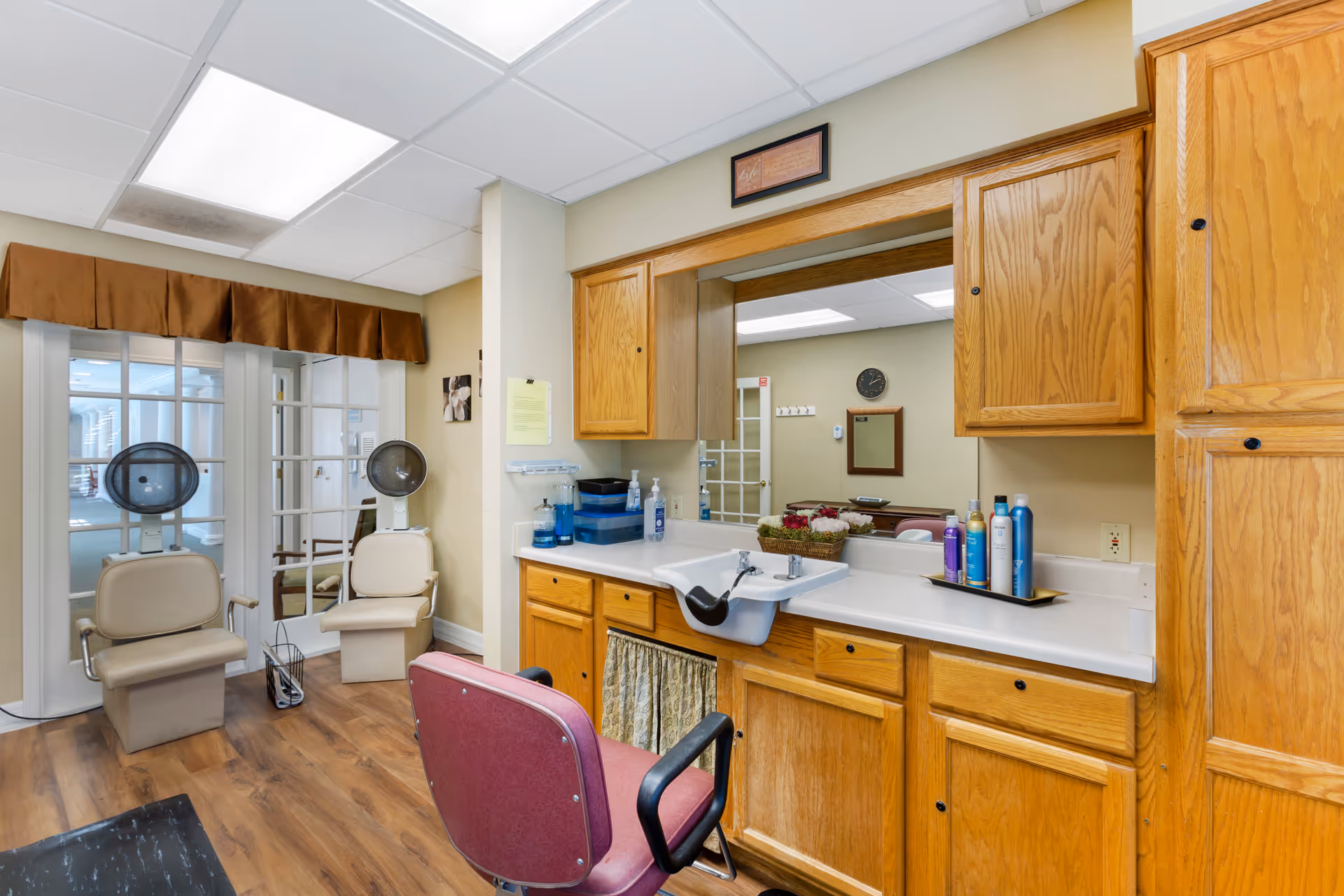 Interior view of a salon area in a senior living facility with wooden cabinets, a large mirror, a white sink, and a red salon chair in front of the sink. Two beige salon chairs with hair dryers are positioned near glass-paneled double doors. The floor is wood, and the walls are painted beige.