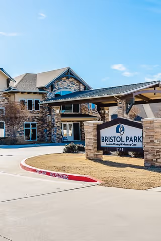 Exterior view of Bristol Park at Eagle Mountain Assisted Living & Memory Care facility showing a stone and brick building with a covered entrance and a sign in front displaying the facility name and address 3141.