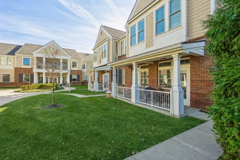 Exterior view of a senior living facility with beige and brick buildings, green lawn, small tree, and a concrete walkway under a partly cloudy sky.