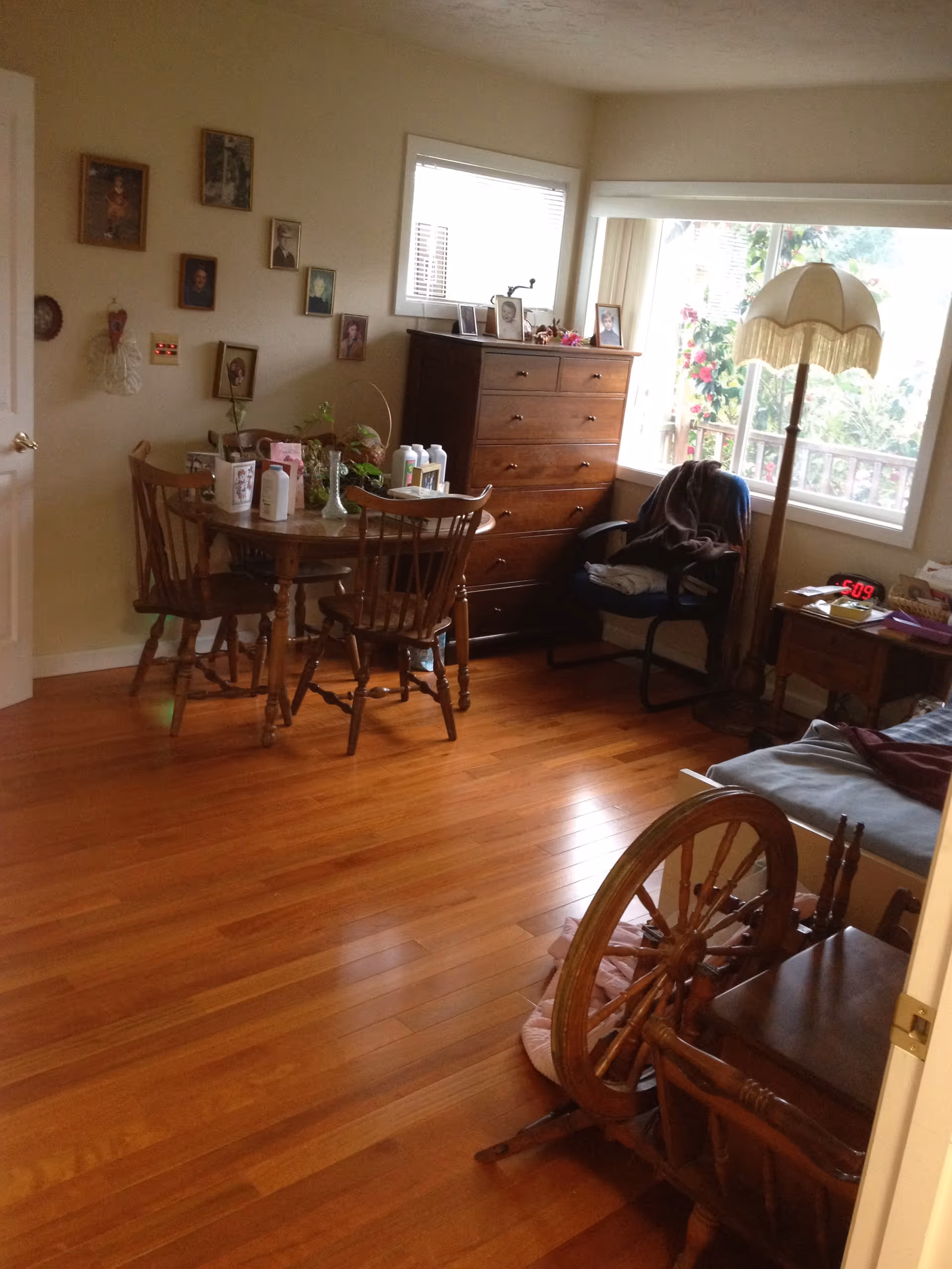A cozy room with wooden flooring featuring a round wooden table with four chairs, a tall wooden dresser, a chair with folded clothes, a vintage floor lamp, and a window with a view of greenery outside. Several framed photos and decorations are on the walls and furniture.
