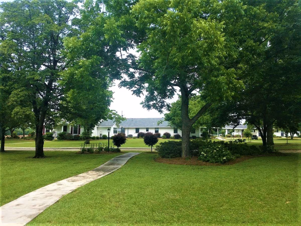 A paved walkway leads through a well-maintained grassy lawn with several large green trees and shrubs. In the background, there are single-story white buildings with dark roofs, likely part of a senior living facility, under a partly cloudy sky.