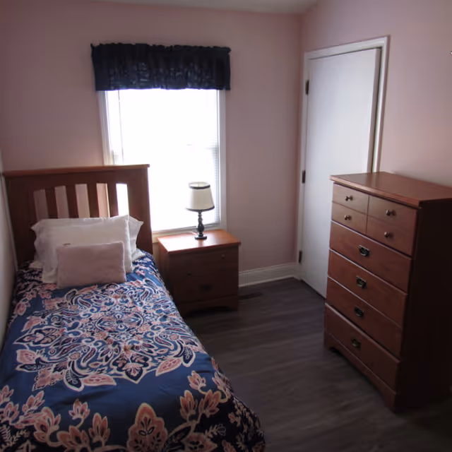 A small bedroom with a single wooden bed covered with a blue and pink patterned bedspread, a white pillow, and a smaller pink pillow. Next to the bed is a wooden nightstand with a small lamp. Across from the bed is a wooden chest of drawers. The room has light pink walls, a window with blinds and a dark valance, and a closed white door.