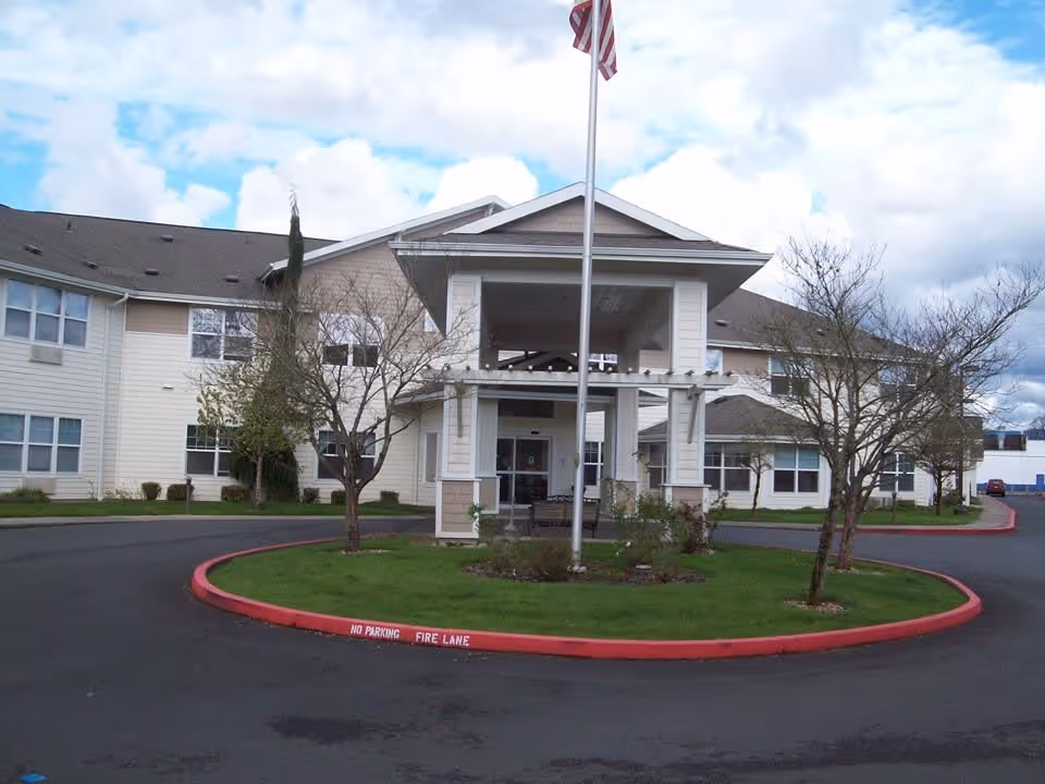Exterior view of Mallard Landing by Cogir, showing the front entrance with a covered driveway, a flagpole with an American flag, and a circular landscaped area with grass and small trees. The building is two stories with beige siding and multiple windows under a cloudy sky.