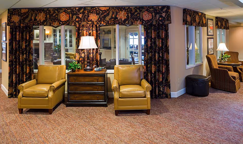 A cozy seating area in a memory care community featuring two mustard yellow leather armchairs separated by a wooden side table with a lamp and a small plant. Behind the chairs are large windows with floral-patterned curtains, revealing another room with additional seating. To the right, there is a small round table with two patterned armchairs and a lamp, all set on a carpeted floor.