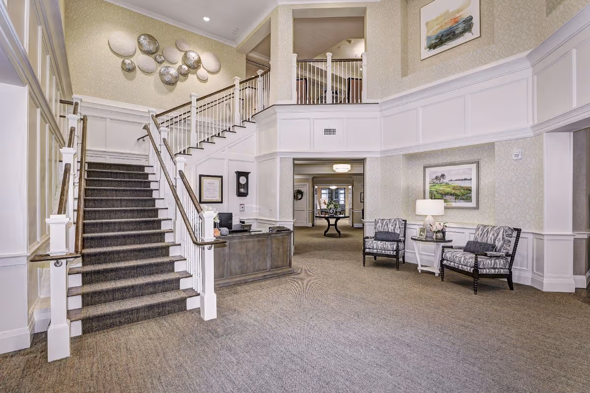 Bright, elegant senior living lobby with a central carpeted staircase, reception desk, and seating area under decorative wall art.