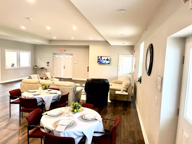 Bright communal dining and lounge area with round tables set for meals and sofas facing a wall-mounted TV.