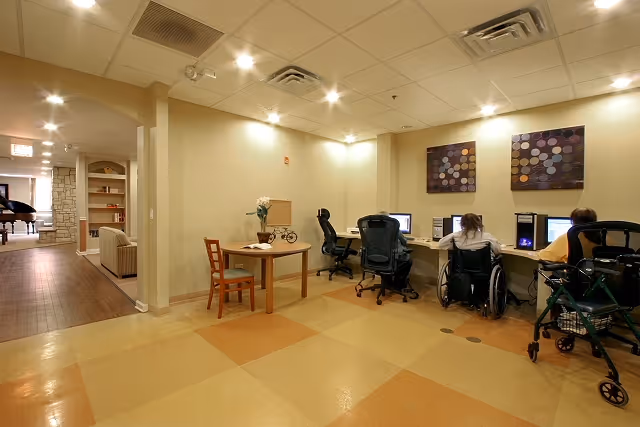 A room with a tiled floor and beige walls featuring a small wooden table with a chair and a decorative bicycle on it. Three people are seated at a long desk using desktop computers; one person is in a wheelchair and another has a walker nearby. The room is well-lit with ceiling lights and has two abstract paintings on the wall. In the background, there is a hallway leading to another room with a piano and seating area.