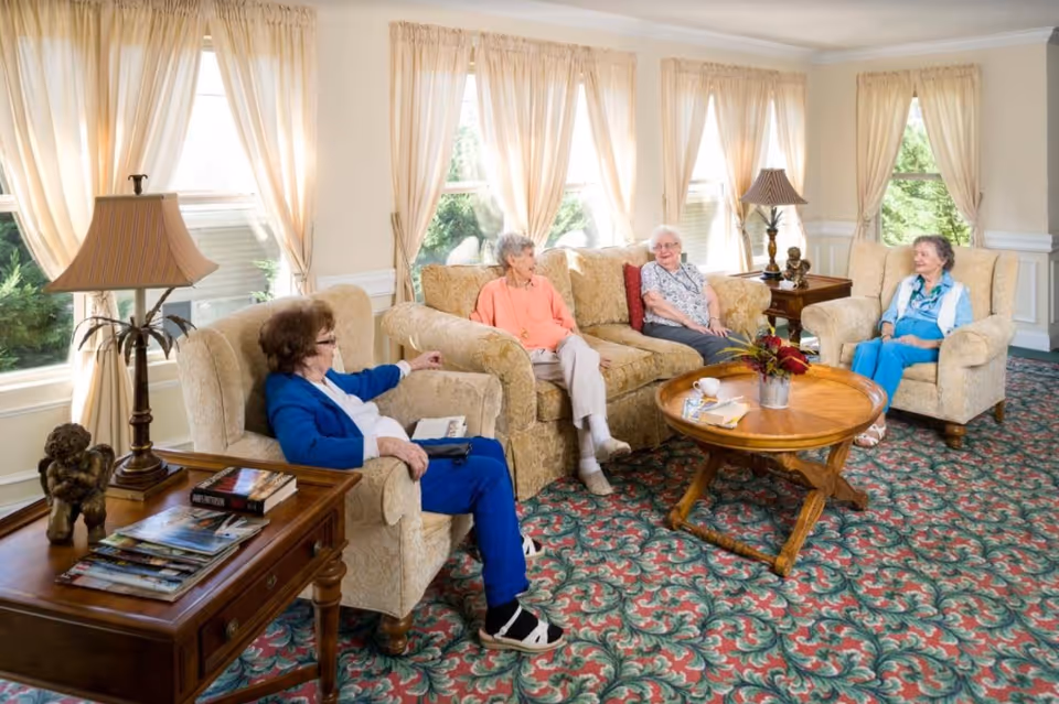 A bright living room with large windows covered by light curtains. Four elderly women are seated on beige upholstered armchairs and a sofa, engaged in conversation. The room features a patterned carpet, wooden side tables with lamps, and a round wooden coffee table with a flower arrangement and some magazines.