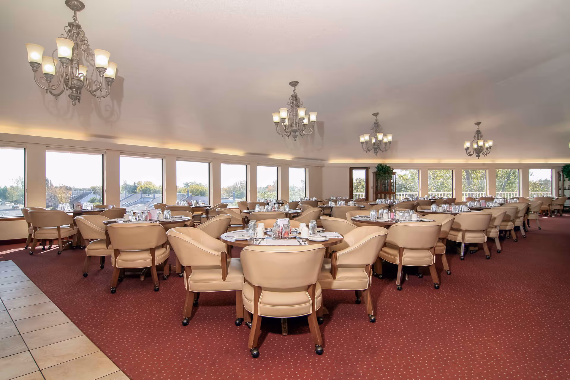 A spacious dining room with multiple round tables set with plates, glasses, and silverware. Each table is surrounded by beige cushioned chairs with wooden legs. The room has large windows letting in natural light and several ornate chandeliers hanging from the ceiling. The floor is carpeted in red with a small tiled area visible on the left side.