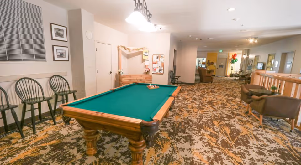 Interior view of a common area in a senior living facility featuring a pool table with balls arranged for a game, several chairs along the wall, a small seating area with two armchairs and a table, and a carpeted floor with a patterned design. The space is well-lit with ceiling lights and decorated with framed pictures and bulletin boards on the walls.