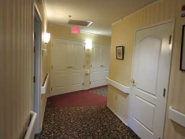 Interior hallway of a senior living facility with patterned carpet, beige striped wallpaper, white doors, handrails along the walls, and wall-mounted lights. There is an exit sign above a set of double doors at the end of the hallway.