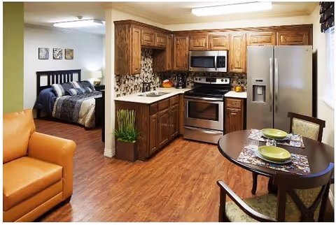 A cozy interior space featuring a kitchen with wooden cabinets, stainless steel appliances including a refrigerator, stove, and microwave. Adjacent to the kitchen is a small round dining table set with two place settings. To the left, there is a partial view of a living area with a tan leather armchair and an open doorway leading to a bedroom with a bed, nightstand, and wall art.