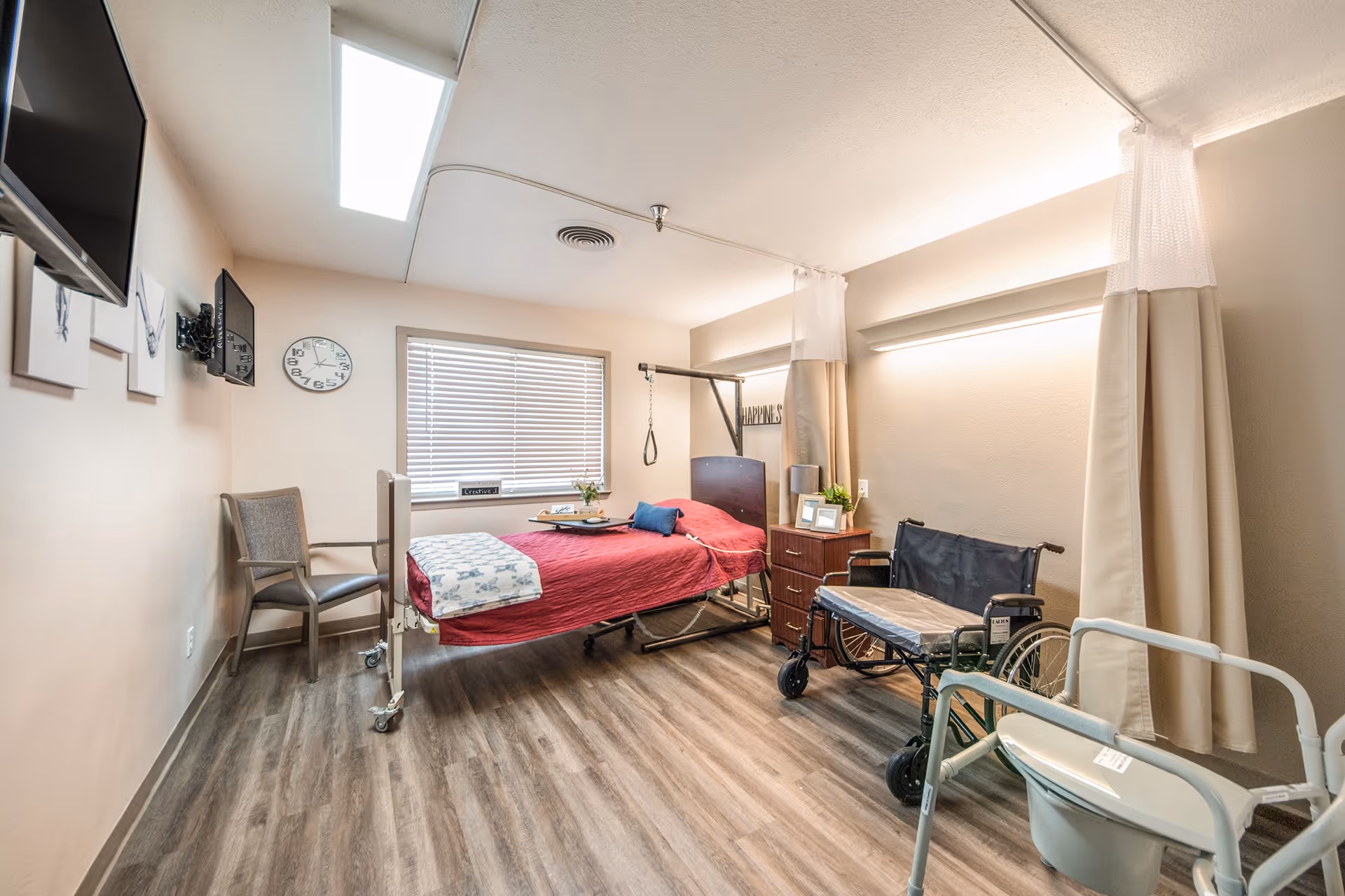 A clean senior care bedroom with a single hospital-style bed, wheelchair, bedside table, and privacy curtains.