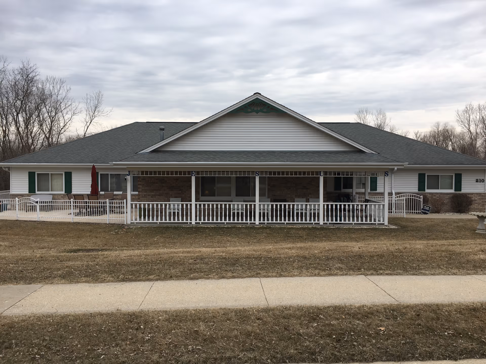 Exterior view of a single-story senior living facility building with a gray roof, white siding, and green shutters. The building has a covered porch with white railings and several chairs. There is a sidewalk and a grassy area in front of the building, and leafless trees are visible in the background under a cloudy sky.