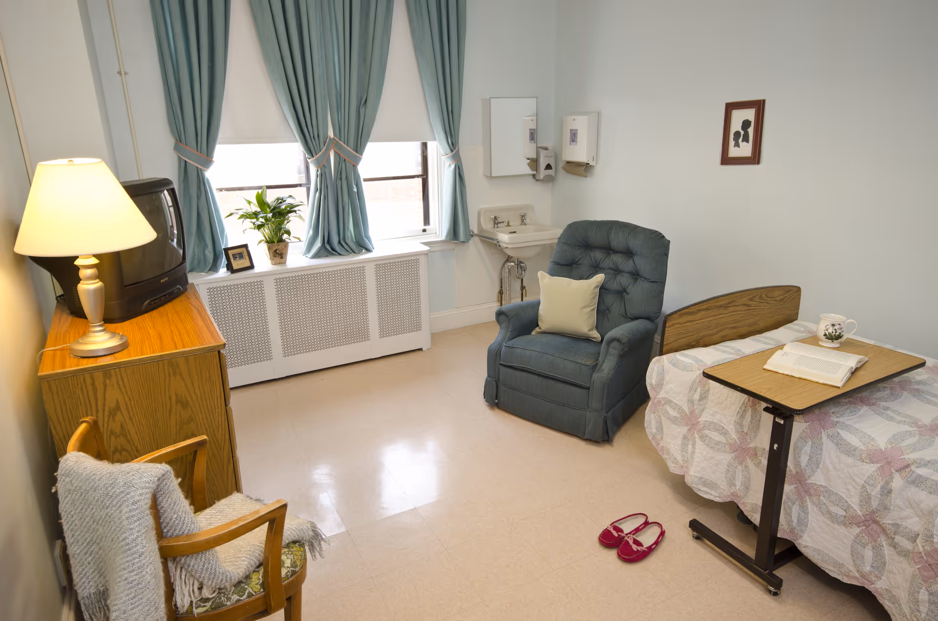 A small, tidy room with a single bed covered with a patterned quilt, a blue recliner chair with a pillow, a wooden side table with a lamp and an old-style TV, a wooden chair with a blanket draped over it, and a window with blue curtains. There is a small sink with a mirror and soap dispenser on the wall, a pair of red slippers on the floor, and a framed silhouette artwork hanging on the wall.