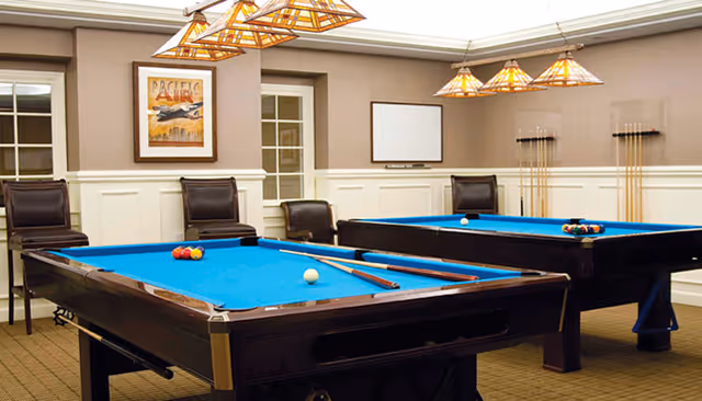 A game room with two pool tables featuring blue felt tops, several pool cues mounted on the wall, and four dark leather chairs. The room has beige walls with white wainscoting, a framed poster on the wall, and pendant lights hanging from the ceiling.
