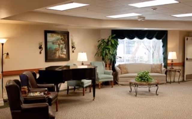 Common sitting area with a grand piano, several armchairs, a sofa, a coffee table and lamps under ceiling lights.