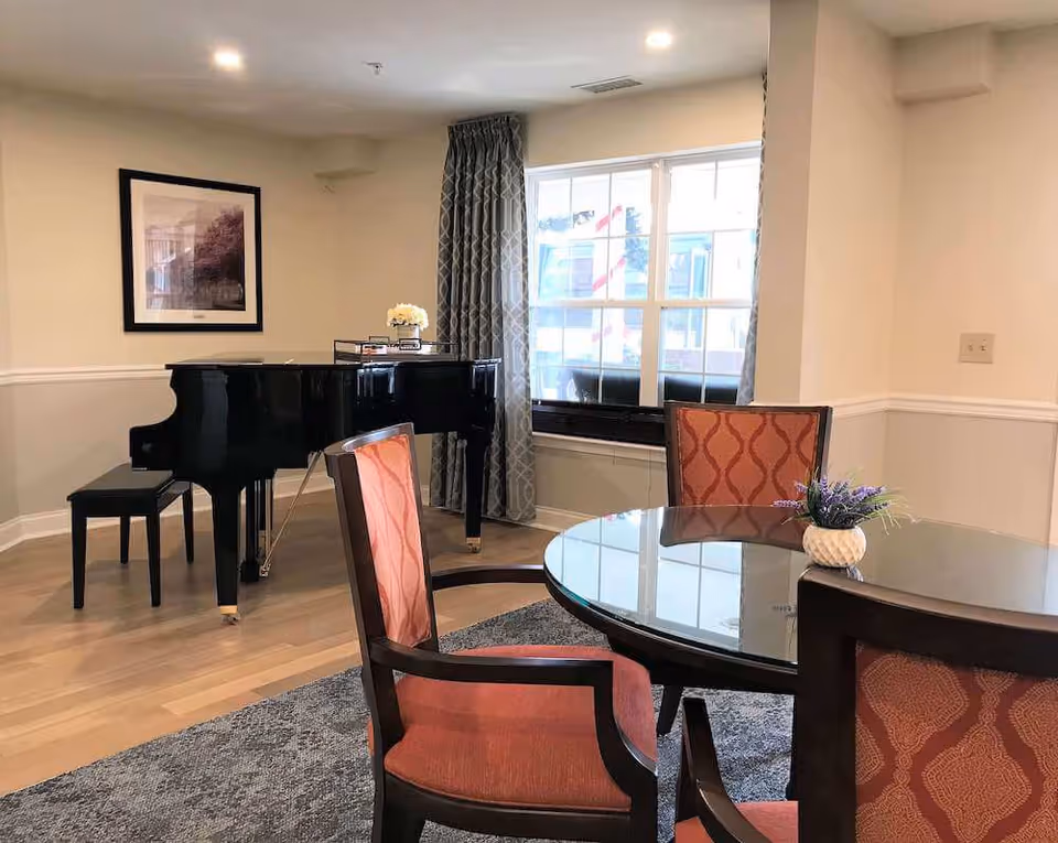 Bright communal dining area with a round glass table and upholstered chairs, a grand piano by the window, and framed art on the wall.