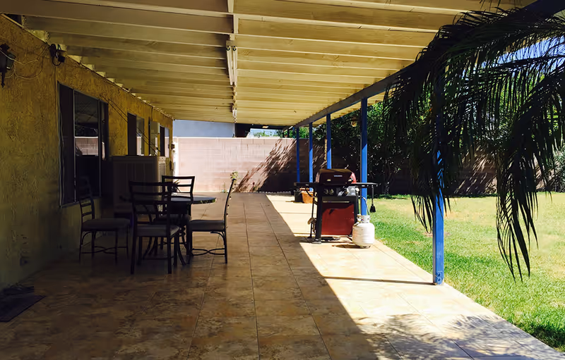 Covered backyard patio with a tiled floor, a dining table and chairs, a gas grill, and an adjacent grassy yard with palm fronds.