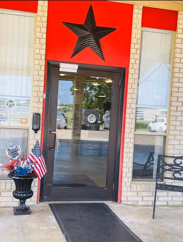 Entrance door of Willow Park Rehabilitation and Healthcare Center with a large black star decoration above the door on a red background. There is a black planter with red, white, and blue decorations and an American flag to the left of the door, and a black metal bench to the right. The door is glass with a metal frame, reflecting parked cars and trees outside.