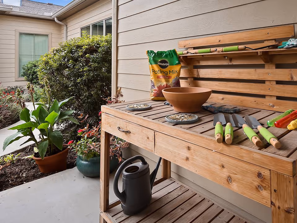 Wooden potting bench on a covered patio with gardening tools, a potting mix bag, potted plants and a watering can next to a building.